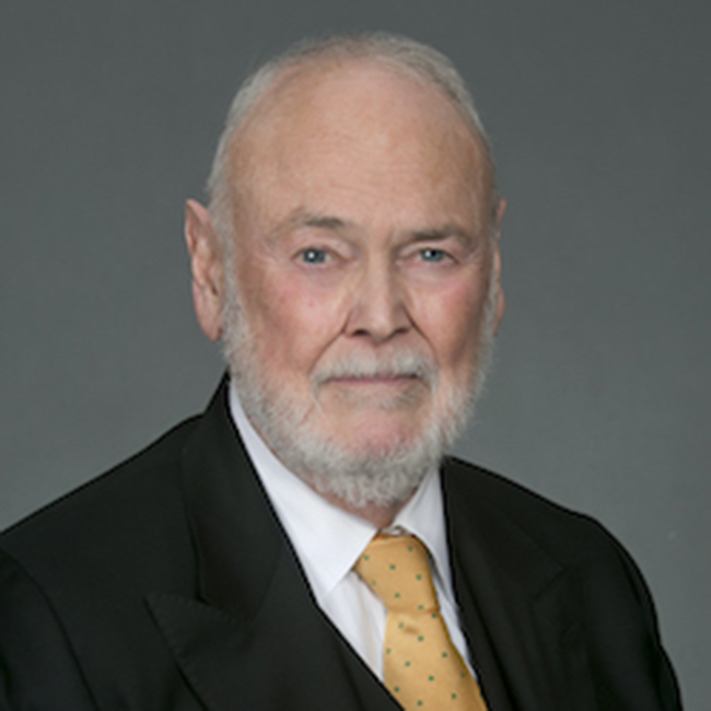 Professional headshot of senior man in black suit and gold tie against gray background.